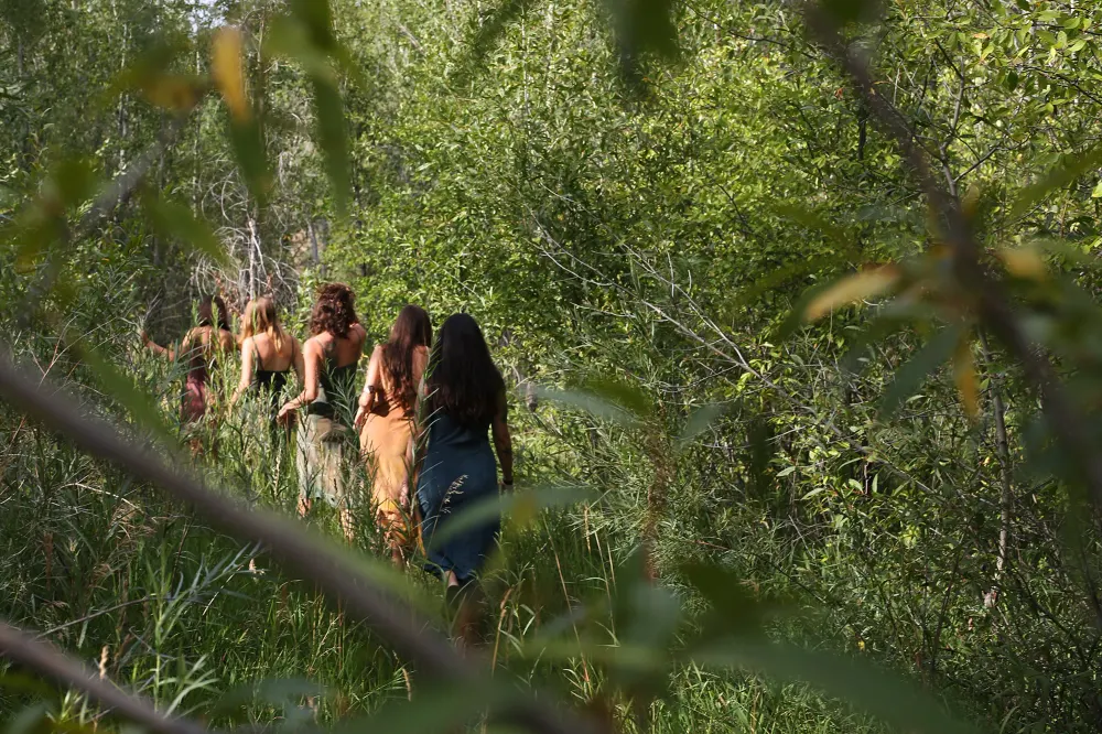 Five women in plant-dyed dresses walking through willows and tall grass
