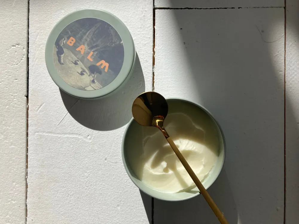 An open tin of balm with a gold spoon, in raking sunlight